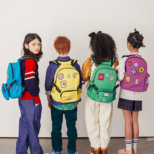 Four kids wearing colourful canvas belt backpacks in blue, yellow, green, and purple, each decorated with fun patches.