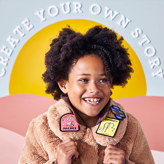 Kid wearing a jacket with "My First Wheels," "Sew-What," and "Maths Wizz" patches attached.