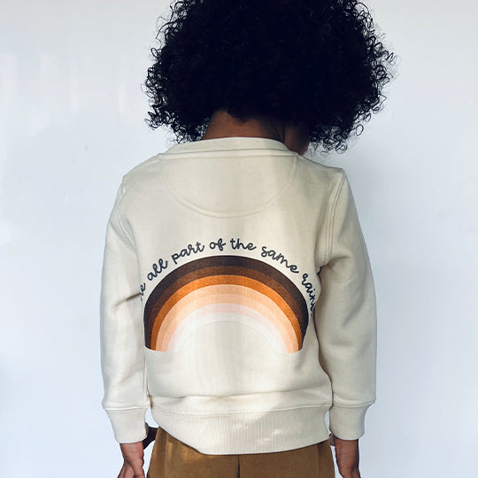 A boy wearing a cream-coloured sweatshirt with a rainbow symbol on the front. Back text reads: "We are all part of the same rainbow