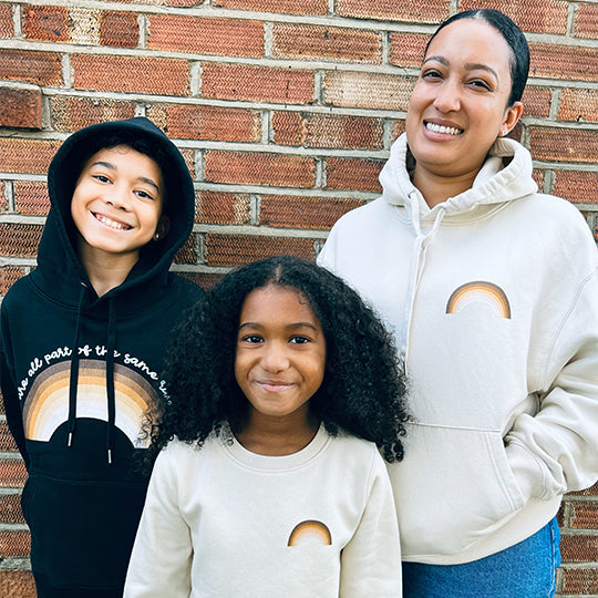 Woman in cream rainbow hoodie, young boy in black hoodie, and girl in cream rainbow sweatshirt made from organic cotton material.