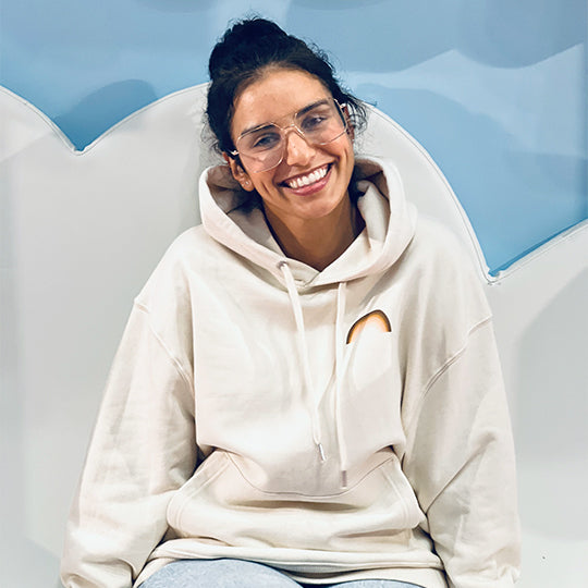 Woman wearing a cream-coloured organic cotton hoodie with "Rainbow" printed on the front. Sustainable, stylish, and comfortable Hoodie.
