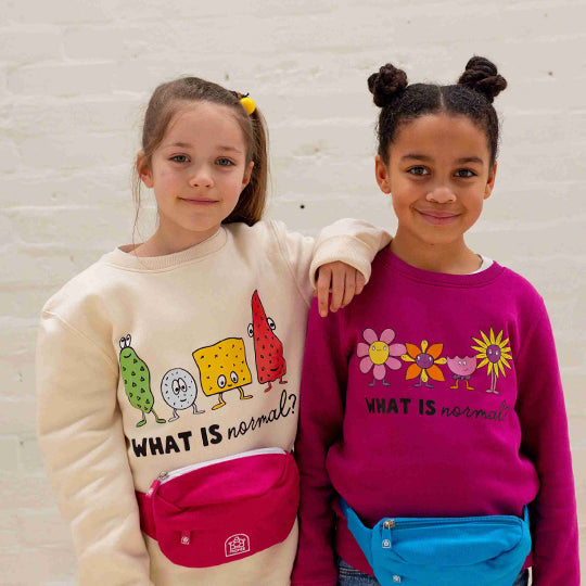 Two young girls wearing organic cotton sweatshirts, one in pink and the other in cream, both printed with "What is normal?" statement.