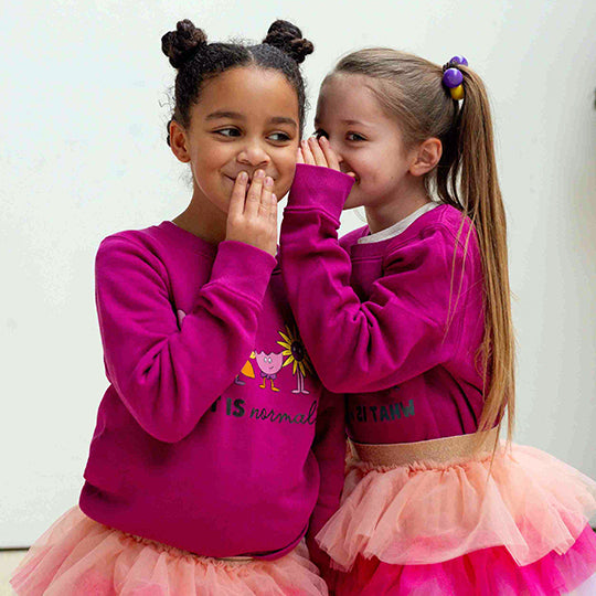 Two young girls smiling in pink cotton sweatshirts with "What is normal?" and cute flower faces printed on them.