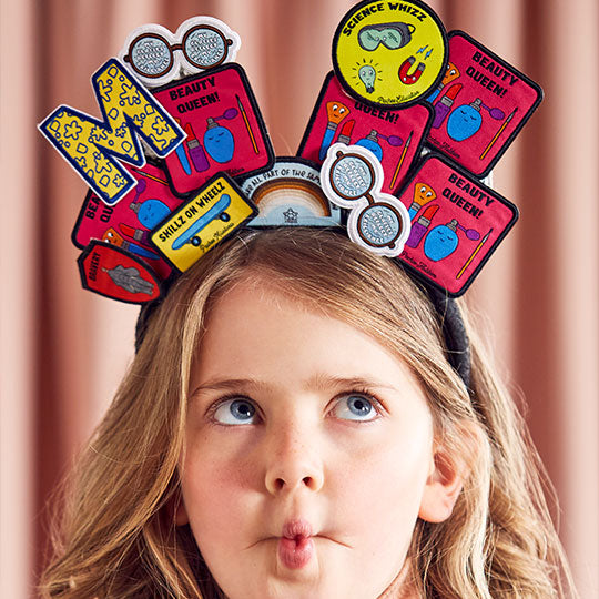 Image of a girl wearing various patches on her head, including "Beauty Queen," "My Glasses," "Science Whizz," "Letter M," and "We Are Part of Same Rainbow'.