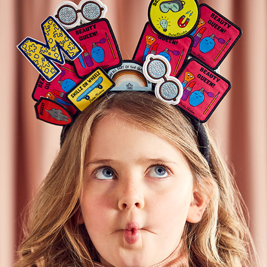 Image of a girl wearing various patches on her head, including "Beauty Queen," "My Glasses," "Science Whizz," "Letter M," and "We Are Part of Same Rainbow'.