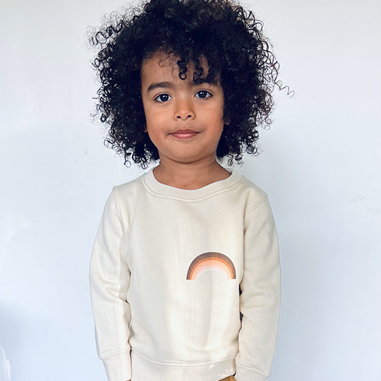 A young boy wearing a cream-coloured sweatshirt featuring a vibrant rainbow design, smiling outdoors on a bright, cheerful day.