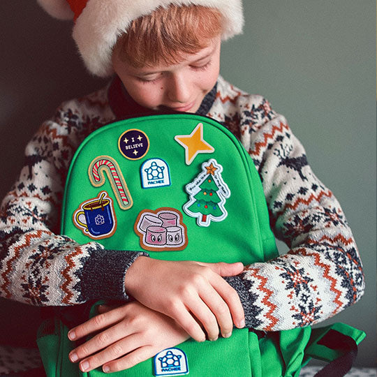 Green backpack with Christmas-themed patches, including a Christmas Star, Tree, 'Coco to my Mallow,' 'I Believe,' Candy Cane Patches.