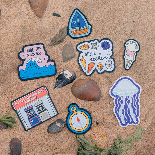 A sealife collection of vibrant patches including Glitter Jellyfish, Shell Seeker, Compass, Arcade, Ride the Waves, Sail Boat, and Ice Cream on beach sand.

