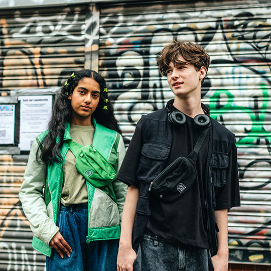 A girl wearing a crossbody green bum bag and a boy wearing a crossbody black bum bag, showcasing stylish and practical accessories.
